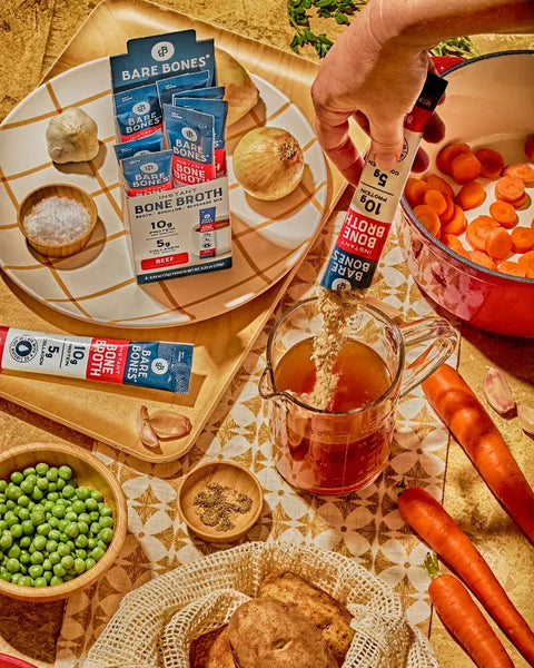 A hand pours powdered bone broth mix into a glass measuring cup filled with broth, surrounded by vegetables, peas, and bone broth packets on a kitchen table.